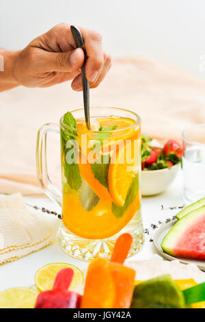 Giovane uomo agitazione un vaso con un drink rinfrescante, con fettine di arancia e menta, collocato su un tavolo accanto a una ciotola con fragole, una piastra con waterm Foto Stock