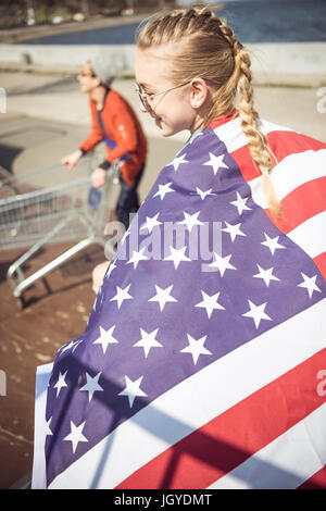 Giovane ragazza bionda in occhiali da sole azienda bandiera americana mentre in piedi sulla rampa di skate park Foto Stock