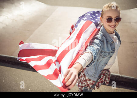 Giovane ragazza bionda in occhiali da sole azienda bandiera americana mentre in piedi sulla rampa di skate park Foto Stock