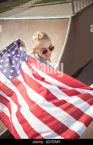 Giovane ragazza bionda in occhiali da sole azienda bandiera americana mentre in piedi sulla rampa di skate park Foto Stock