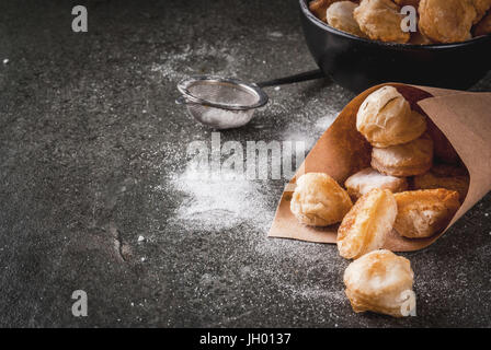 In casa la cottura, sfogliatine. Cibo alla moda. Cronuts popcorn, puff ciambelle fori in una ciotola nero e un sacco di carta, con zucchero a velo. Su una pietra scura Foto Stock