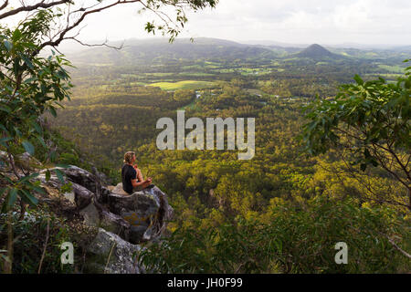 Un giovane uomo si siede tranquillamente, meditando sulla cima di una montagna con ampie vedute vicino a Noosa teste, Australia. Foto Stock