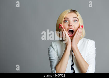 Sorpreso bionda con le mani sul viso sul vuoto lo sfondo grigio Foto Stock