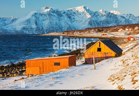 Tipiche baite in legno chiamato Rorbu circondata dalle onde del mare freddo e vette innevate, Djupvik, Alpi Lyngen, Troms, Norvegia Foto Stock