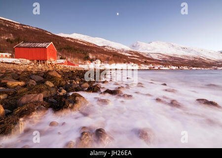 Onde del mare freddo che si infrangono sulle rocce e tipiche baite in legno chiamato Rorbu, Djupvik, Alpi Lyngen, Troms, Norvegia e Scandinavia Foto Stock