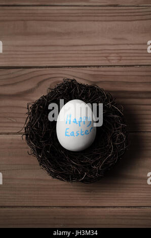 Concetto di pasqua. Tettuccio di colpo di un falso uovo bianco in un nido sul tavolato di legno. Il messaggio 'Felice Pasqua' è scritto sull'uovo in blu. Tonica per un Foto Stock