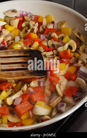 La frittura o sauteing process. Una miscela di verdure fresche in una coppa in ceramica sul piano di cottura della cucina con una spatola di legno. Vapore ascendente visibile nella parte anteriore del pan. Foto Stock