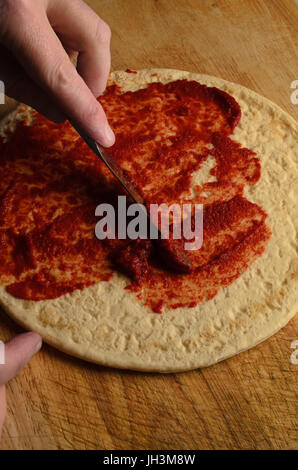 Un maschio di spargimento a mano la salsa di pomodoro sulla base per pizza con il coltello su un vecchio, graffiato e indossato tagliere di legno. Luce oscura. Foto Stock