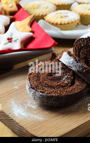 Natale al cioccolato Yule Log su pannello di legno decorato con biscotti e torte di carne macinata in background. Foto Stock