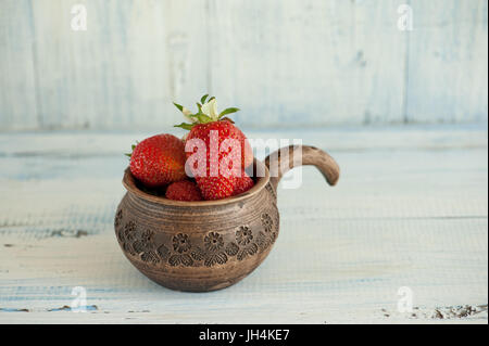 Rosso fragole mature, risiede in un marrone pentola di creta con una penna su un blu sullo sfondo di legno. Il telaio orizzontale. Foto Stock