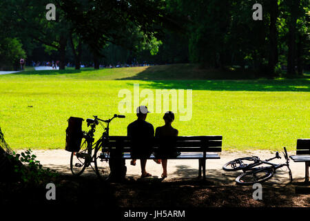 Retroilluminato a coppia giovane con push bike seduta su una panchina nel parco, Englischer Garten Monaco di Baviera, Germania Foto Stock