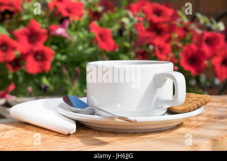 Tazza di caffè servito su un tavolo di legno con un tovagliolo bianco, cucchiaio e biscotto con una sfocata sfondo floreale Foto Stock