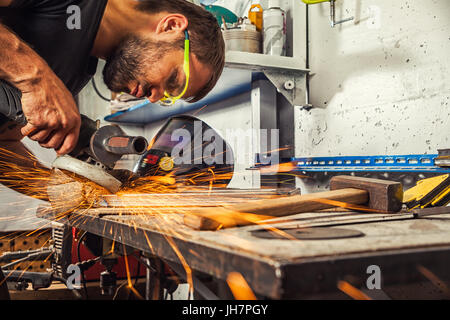 Close-up sui lati volare scintille dalla smerigliatrice angolare. giovane saldatore brunette in black t-shirt e occhiali di grind metal smerigliatrice angolare in officina Foto Stock