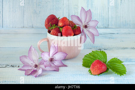 Fragole mature in una coppa in ceramica di colore rosa . Fiori di colore rosa di clematis. Un fondo azzurro. Il telaio orizzontale. Foto Stock