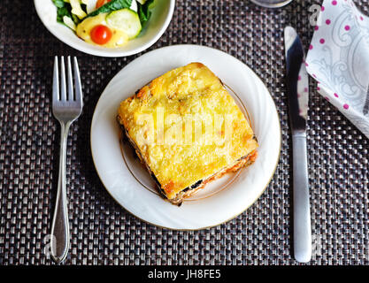 Un pezzo di lasagna di melanzane su un piatto bianco con coltello, forchetta e insalata (sopra) Foto Stock