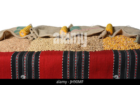 Il farro, soia e chicchi di grano e di chicchi di mais versata da sacchi di iuta che giacciono su un kilim. Le pannocchie sono anche visibili. Su sfondo bianco. Foto Stock