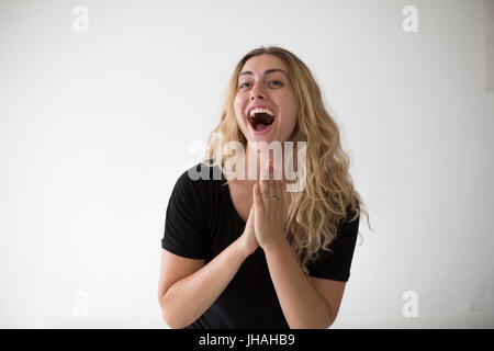 Giovane donna bionda tifo e battendo le mani in emozione e felicità contro uno sfondo bianco in luce naturale del giorno. Una bella millenial. Foto Stock