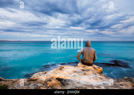 Un uomo si siede sul bordo di un robusto roccia calcarea e si affaccia su un oceano tempestoso vista in Sud Australia. Foto Stock