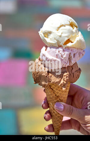 Cioccolato, vaniglia e berry ice cream scoop nel cono di cialda detenute da mano femminile contro lo sfondo colorato Foto Stock