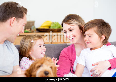Immagine ravvicinata di una famiglia felice seduti insieme sul divano nel soggiorno di casa propria. Essi hanno il cane con loro e stanno parlando e sorridente Foto Stock