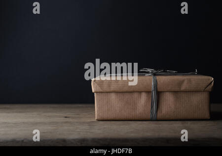 Una confezione regalo, in marrone normale avvolgimento della carta con il grigio scuro rafia nastro su tavola in legno di quercia, con lavagna background fornendo copia spazio. Foto Stock