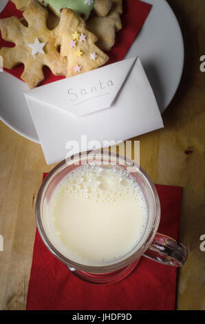 La notte di Natale in scena con una piastra di decorate biscotti) e un bicchiere di latte con busta indirizzata a Santa su un tavolo di legno. Foto Stock