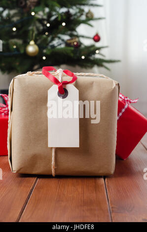 Casa natale in scena con la carta marrone string legato parcel, tag vuoto e nastro rosso sul tavolato di legno con confezioni regalo, decorate albero e tenda in bac Foto Stock