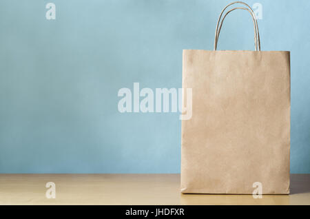 Blank carta marrone borsa con manici per lo shopping, rivolto verso la parte anteriore sul lato destro di una luce di impiallacciatura di legno con tavolo blu pallido sfondo parete stabilito Foto Stock