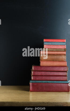 Una pila di vecchi, utilizzati libri di testo impilati in una pila su una scrivania in legno di fronte a una lavagna. Copia spazio a sinistra e sopra. Foto Stock