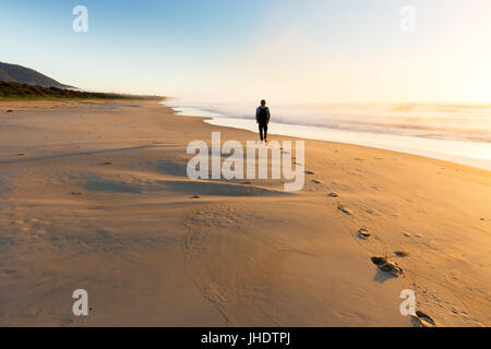 Una persona che cammina su una bellissima spiaggia lascia una scia di impronte come il sorgere del sole illumina la nebbia che sorgono dal mare nella luce dorata. Foto Stock