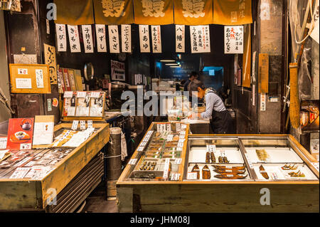 Mercato Nishiki, Kyoto: un pescivendolo negozio vendita varietà di pesce cucinato in molti modi. Un cliente sguardi intorno come il suo ordine è stato avvolto ordinatamente. Foto Stock