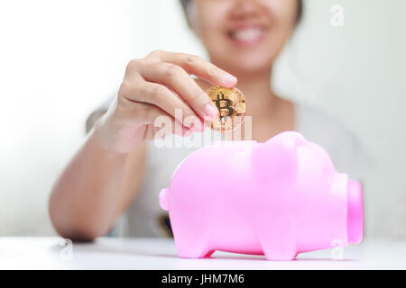 Immagine ravvicinata sorriso donna mettendo bitcoin oro al rosa salvadanaio, profondità di campo, selezionare la messa a fuoco a portata di mano Foto Stock
