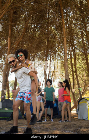 Uomo sorridente piggybacking donna con amici in background in campeggio Foto Stock