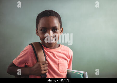 Close up ritratto di ragazzo con zaino e libri contro greenboard nella scuola Foto Stock