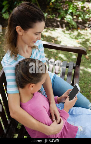 Elevato angolo di visione della donna che guarda la figlia utilizzando il telefono cellulare mentre è seduto sulla panca di legno nel cortile posteriore Foto Stock