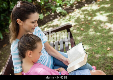 Inclinare il colpo di madre romanzo di lettura alla figlia sul banco di legno in giardino Foto Stock