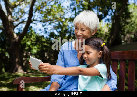 Sorridente ragazza tenendo selfie con la nonna mentre è seduto sul banco a backyard Foto Stock