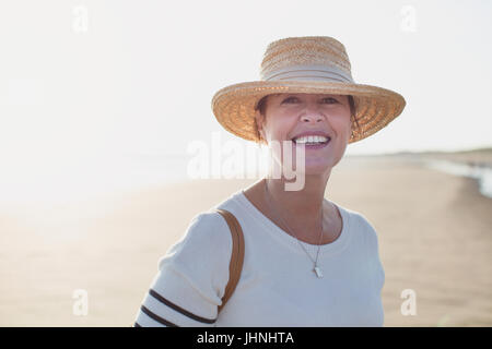 Ritratto sorridente donna matura indossando cappello di paglia sulla soleggiata spiaggia di estate Foto Stock