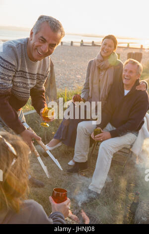 Sorridente coppie mature di bere il vino e la cottura alla brace sulla spiaggia al tramonto Foto Stock