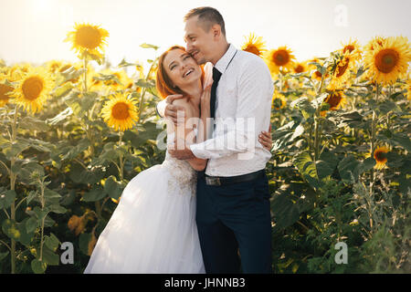 Sposa redhead in matrimonio abiti bianchi e lo sposo whit camicia e cravatta abbracciando permanente nel campo di girasoli. Con sole estate Foto Stock