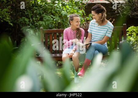 Felice madre e figlia guardando ogni altro mentre è seduto con il romanzo sulla panca di legno nel cortile posteriore Foto Stock