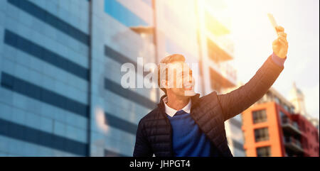 Uomo sorridente prendendo un seflie contro la riflessione sul palazzo di vetro Foto Stock