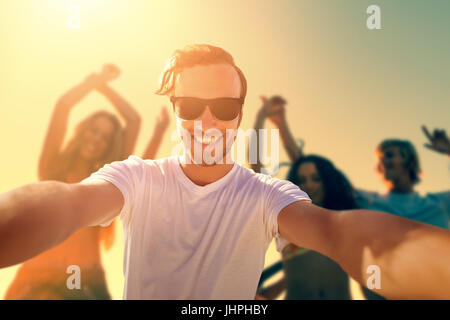 Uomo sorridente prendendo un seflie contro il gruppo di amici Ballare in spiaggia durante il tramonto Foto Stock