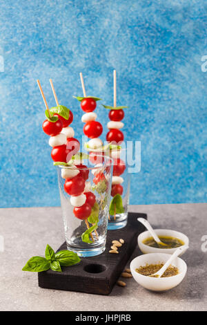 Appetitosi kebab vegetariani da pomodori ciliegia e mini mozzarella in due tipi di basilico e origano sugo come uno snack. Su uno sfondo blu Foto Stock