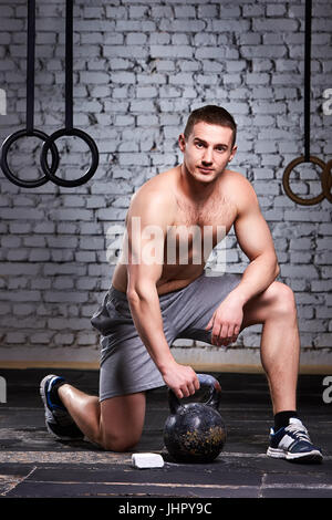 Giovane atleta accovacciato su una gamba e azienda kettlebell contro un muro di mattoni in croce montare palestra. Foto Stock