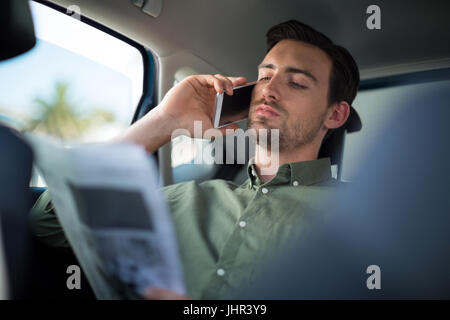 Uomo che parla al telefono cellulare durante la lettura del giornale in auto Foto Stock