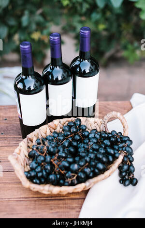 Tre bottiglie di vino rosso e il grappolo di uva labruscan in una piastra wattled su un tavolo di legno, all'aperto. aperitivo a una festa di nozze. Foto Stock