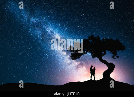 La Via Lattea con abbracciando giovane sotto l'albero sulla collina. Paesaggio con notte cielo stellato e silhouette di standing l uomo e la donna. La Via Lattea con voi Foto Stock
