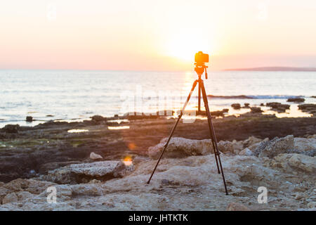 Fotocamera con treppiede oltre il sole che sorge nei pressi della spiaggia Foto Stock