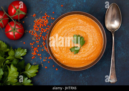 Purea di zuppa di lenticchie rosse in una ciotola marrone, pomodori freschi, prezzemolo su uno sfondo blu. vista da sopra Foto Stock